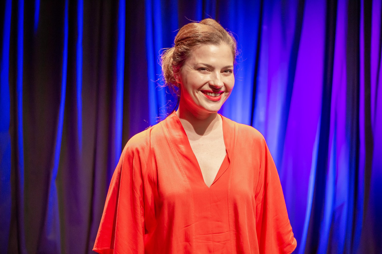 Foto colorida de Elen Londero. Ela veste uma blusa laranja, está com o cabelo preso e ao fundo tem uma cortina iluminada na cor azul