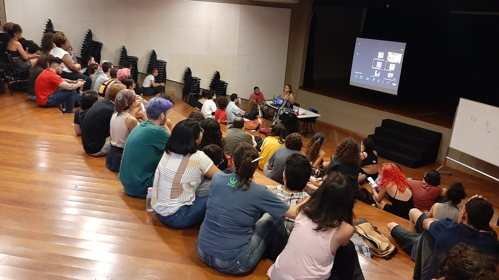 Foto colorida mostrando os estudantes da SP Escola de Teatro sentados no tablado do auditório da sede Brás
