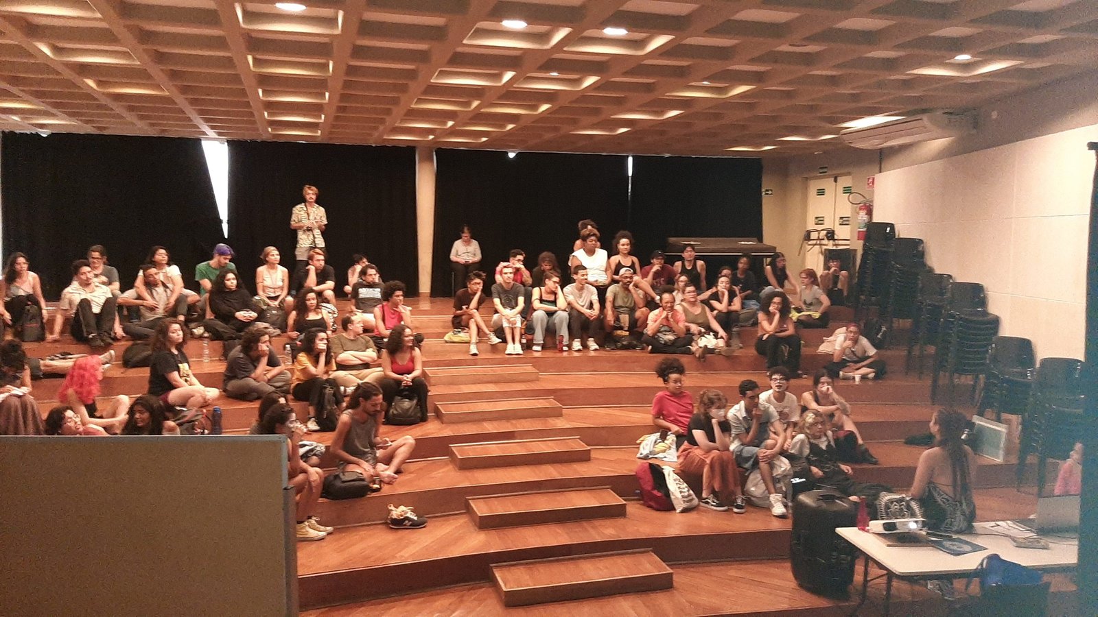 Foto colorida mostrando os estudantes da SP Escola de Teatro sentados no tablado do auditório da sede Brás