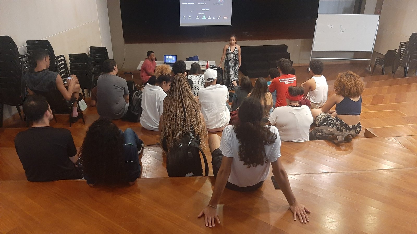 Foto colorida mostrando os estudantes da SP Escola de Teatro sentados no tablado do auditório da sede Brás