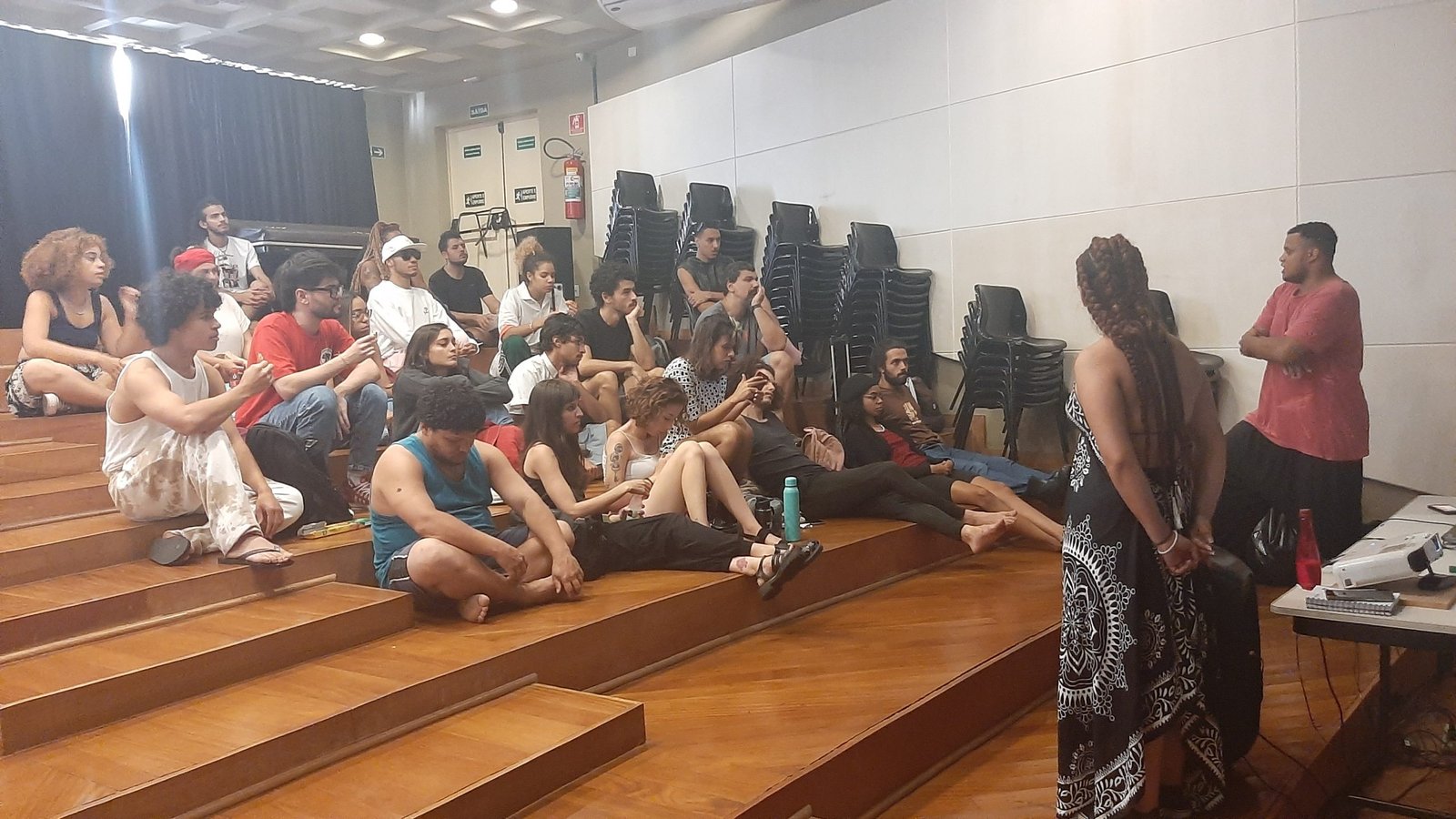 Foto colorida mostrando os estudantes da SP Escola de Teatro sentados no tablado do auditório da sede Brás