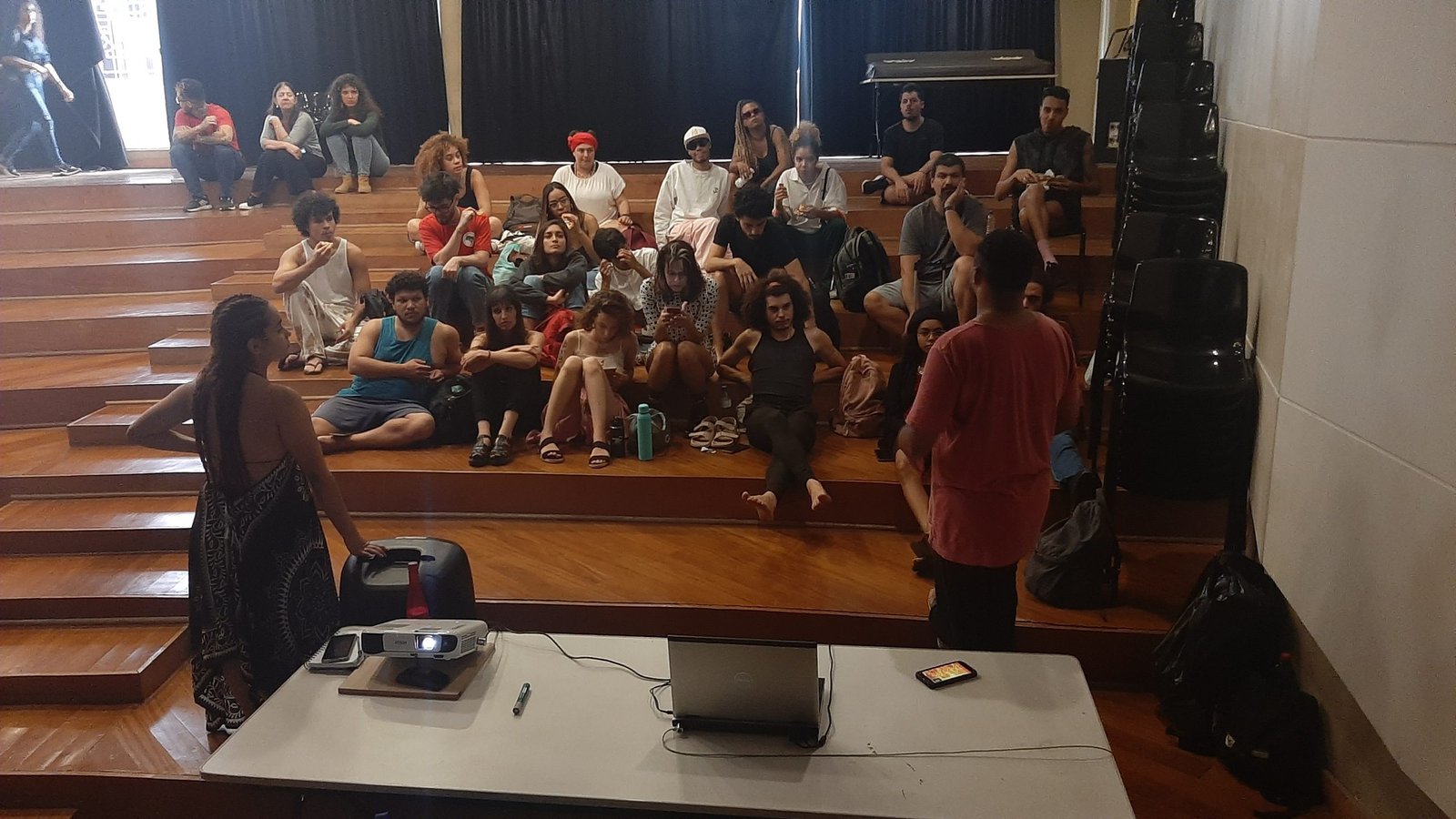 Foto colorida mostrando os estudantes da SP Escola de Teatro sentados no tablado do auditório da sede Brás