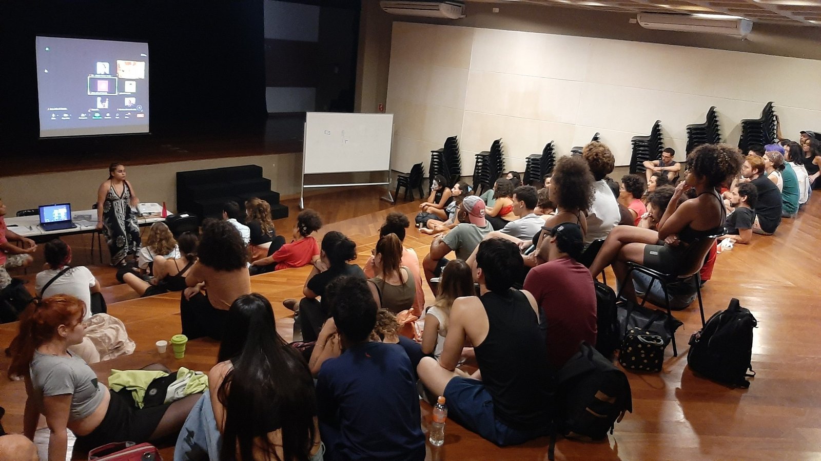 Foto colorida mostrando os estudantes da SP Escola de Teatro sentados no tablado do auditório da sede Brás