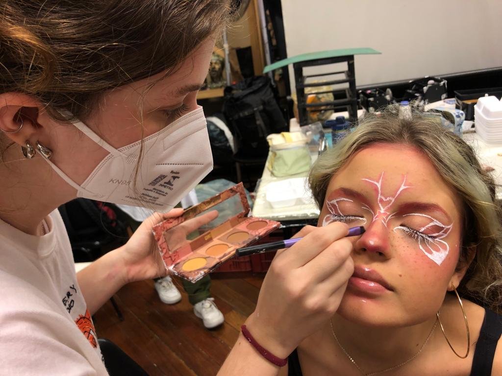 Foto colorida mostrando uma estudante fazendo maquiagem em outra estudante. A maquiagem tem as cores rosa e branca e possuem um desenho de borboleta na região dos olhos. 