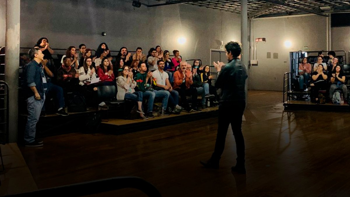 Foto colorida mostrando Pedro Leão de costas aplaudindo o plateia, a qual está sentada à sua frente num tablado com cadeiras e também ao seu lado direito num mesmo tipo de tablado.
