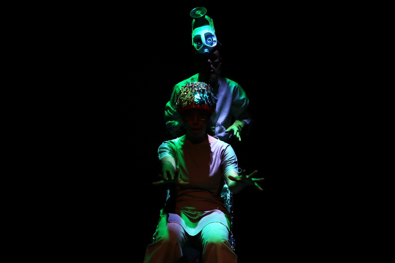 Foto colorida com luz escura mostrando dois estudantes da SP Escola de Teatro, um está sentado e outro atrás dele em pé, em experimento cênico