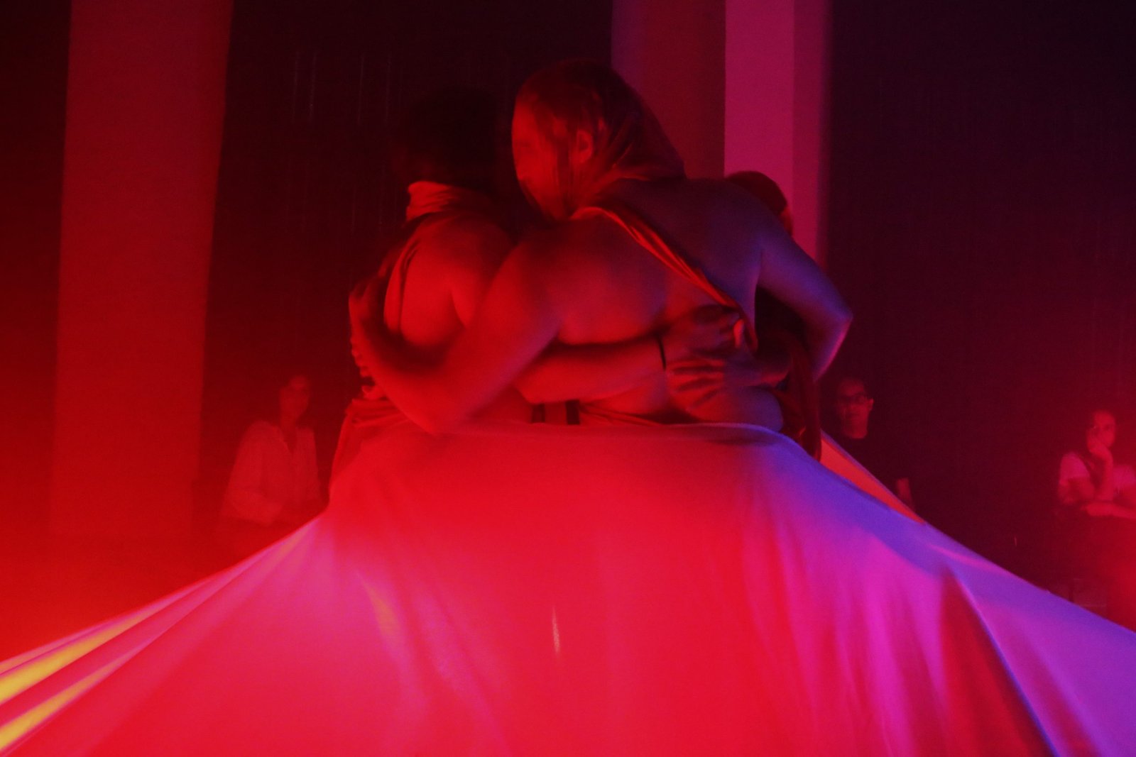 Foto colorida com luz vermelha mostrando estudantes da SP Escola de Teatro abraçados em experimento cênico