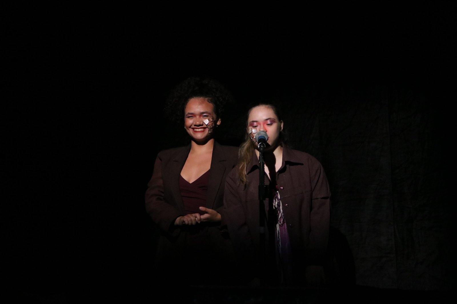 Foto colorida com fundo preto mostrando duas estudantes da SP Escola de Teatro falando no microfone em experimento cênico