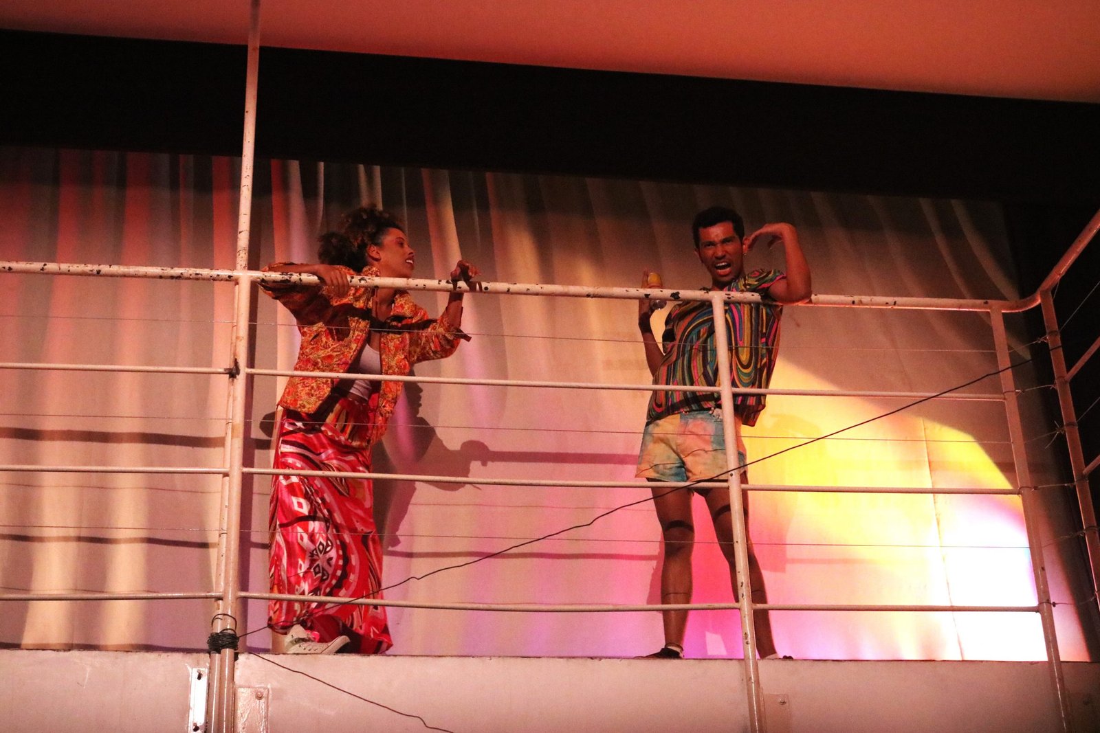 Foto colorida mostrando dois estudantes da SP Escola de Teatro se apoiando na grade da parte superior da sala da sede Roosevelt em experimento cênico