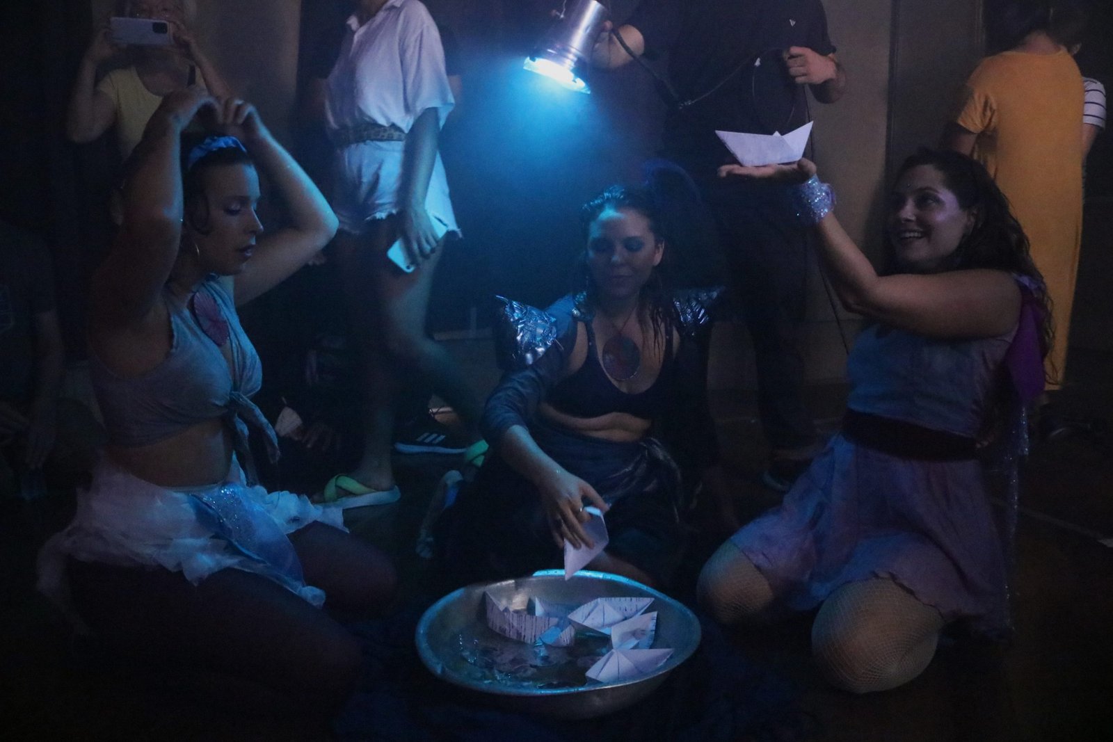 Foto com luz azul mostrando três estudantes da SP Escola de Teatro ajoelhadas no chão com barquinhos de papel em suas mãos em experimento cênico