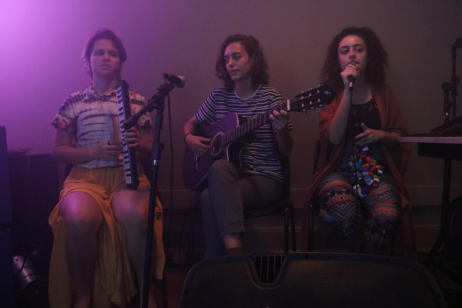 Foto colorida mostrando três estudantes da SP Escola de Teatro, uma tocando escaleta, a outra violão e a terceira cantando em experimento cênico