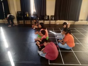 Estudante Alicia e as crianças bolivianas sentadas em atividade da oficina oferecida em uma das salas da sede Brás da SP Escola de Teatro