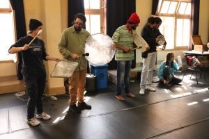 Fotografia colorida mostra estudantes e Barulho Max tocando instrumentos criados em aula por eles mesmo