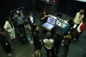 Fotografia colorida da turma de sonoplastia com Allyne Cassini. A orientadora está mostrando uma mesa de som