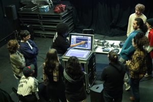 Fotografia colorida da turma de sonoplastia com Allyne Cassini. A orientadora está mostrando uma mesa de som