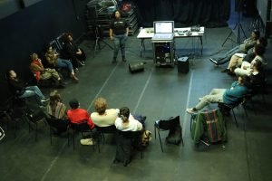 Fotografia colorida da turma de sonoplastia com Allyne Cassini. Todos estão sentados em meia lua