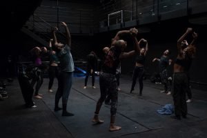 Fotografia colorida mostra aula do curso de "D´água movimento do corpo". Todos estão em pé realizando prática de movimentos corporais.