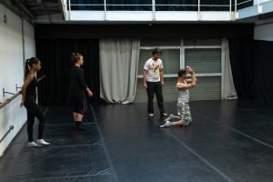 Fotografia colorida mostra aula do curso de "Contato Improvisação em Cena". Um dos participantes está ajoelhado e o orientador e outras duas participantes estão em pé olhando o movimento.