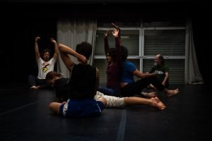 Fotografia colorida mostra aula do curso de "Contato Improvisação em Cena". Todos estão sentados no chão