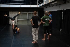 Fotografia colorida mostra aula do curso de "Contato Improvisação em Cena". Enquanto o orientador faz um espacate de ponta cabeça, os participantes observam de pé o movimento.