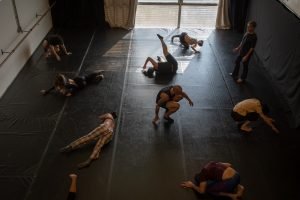 Fotografia colorida mostra aula do curso de "Dança Contemporânea". Todos estão fazendo movimentos diversos no chão