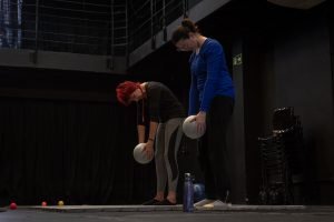 Fotografia colorida mostra aula do curso de "Pilates". A orientadora e a participantes estão em pé com a cabeça baixa segurando uma bola