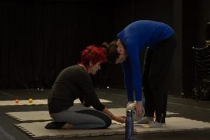 Fotografia colorida mostra aula do curso de "Pilates". A participante está em pé com o corpo abaixado segurando a bola no tatame e a orientadora ajoelhada no tatame.