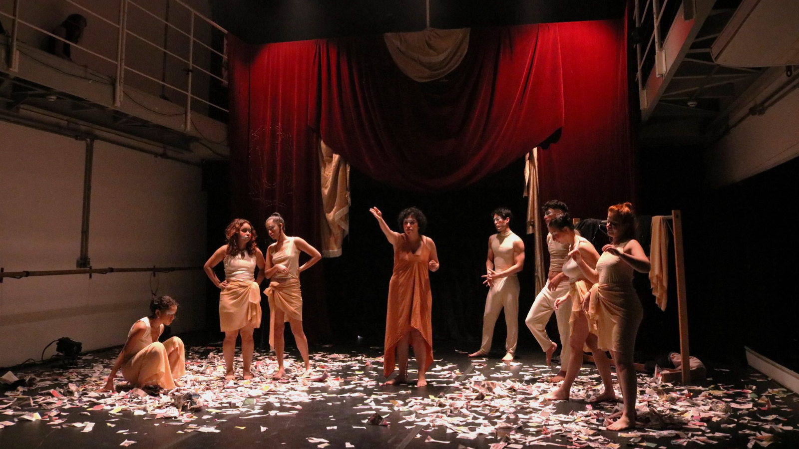 Fotografia colorida mostrando estudantes em experimento cênico em uma das salas da sede Roosevelt. O fundo do cenário é composto por uma cortina vermelha