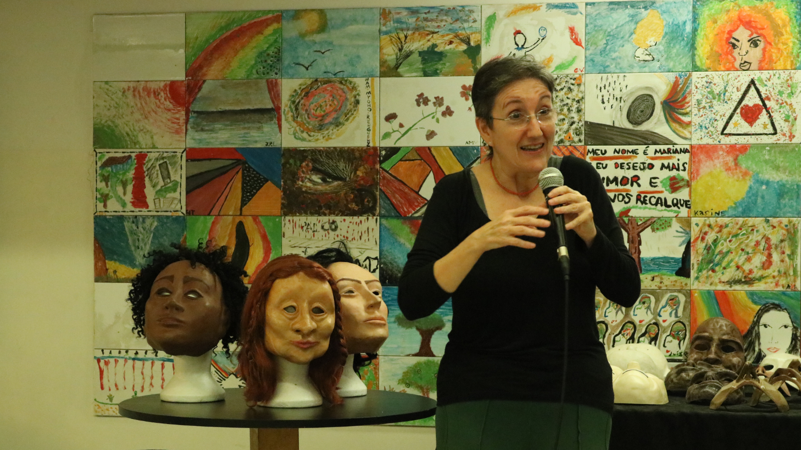 Fotografia colorida mostrando Tiche Vianna segurando um microfone, no hall da sede Roosevelt da SP Escola de Teatro. Ao seu lado esquerdo tem uma mesa redonda com três máscaras.